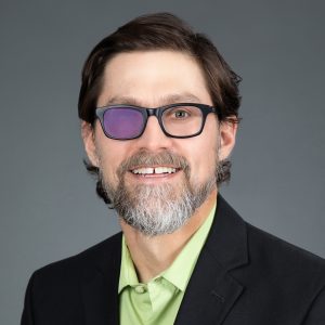 White man with brown hair and a neat beard smiles openly. He wears black frame glasses with a vibrant purple right lens and clear left lens and a black blazer over a pastel green collared shirt. He is against an even professional gray background.