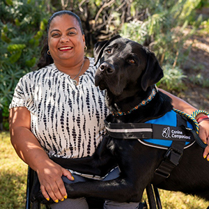 Brown woman with dark hair pulled back in a pony tail, smiling. She is sitting in a wheel chair in a white shirt with black pattern . A black service dog has his two front legs across her lap, the dogs head comes up to her shoulder. She and the dog are against a background of green trees and grass.   