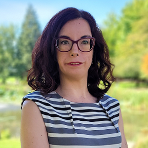 White woman with dark shoulder length hair, looking straight ahead. she wears glasses and a stripped blue, white and black top. She is against a background with trees and a blue sky.