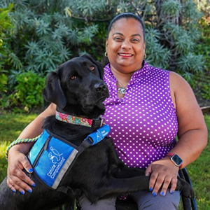 Brown woman with dark hair pulled back in a pony tail, smiling. She is sitting in a wheel chair in a purple shirt with white polka dots. A black service dog has his two front legs across her lap, the dogs head comes up to her shoulder. She and the dog are against a background of green trees and grass.   