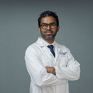 Indian man with glasses wearing a white medical jacket with a blue tie, standing with his arms crossed against a professional gray background. 