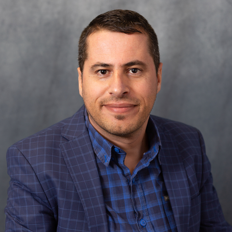 Man with brown hair looking straight at the camera wearing a blue blazer against a grey background. 